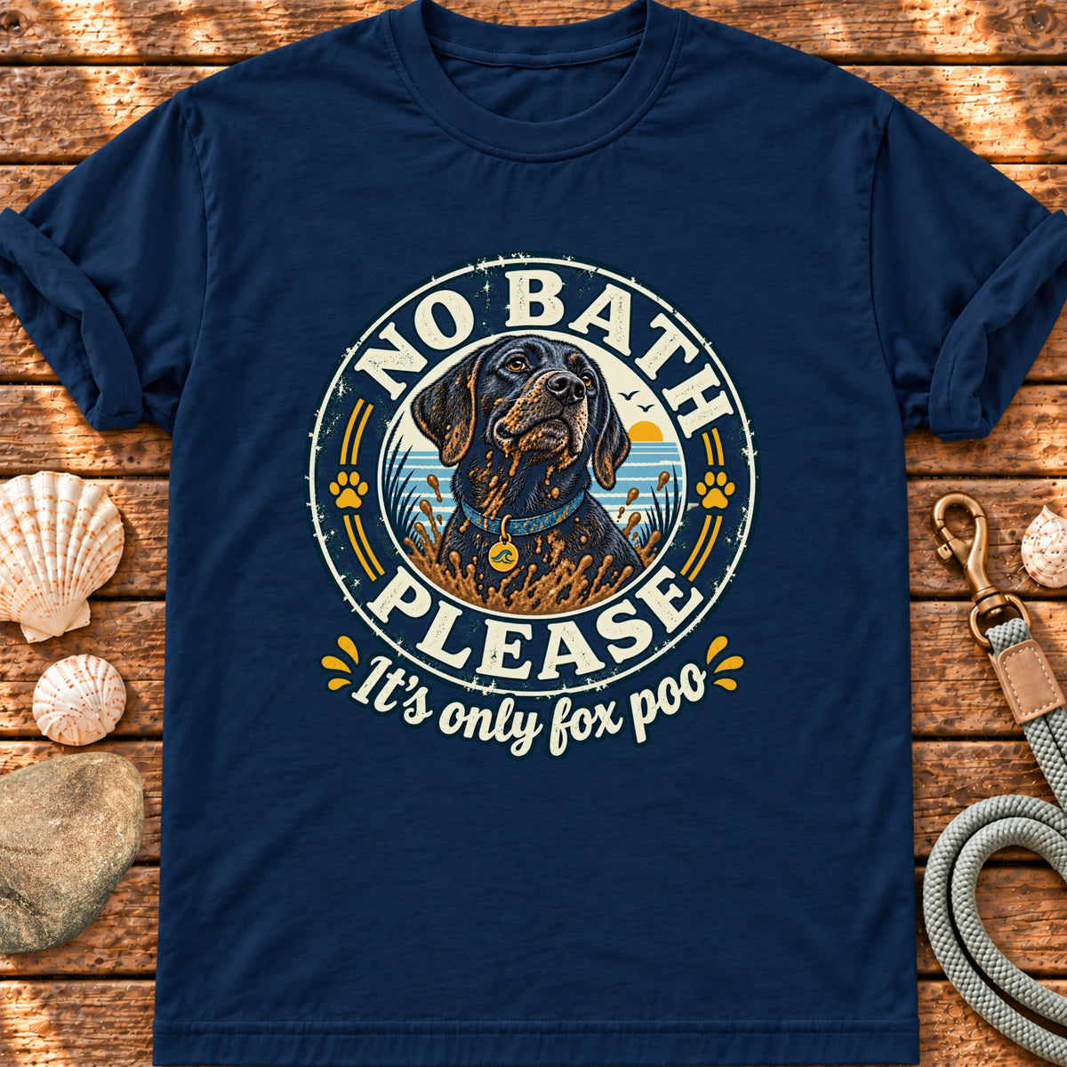 A navy blue short-sleeve t-shirt with a circular graphic of a black dog and the text 'NO BATH PLEASE It's only fox poo' in white and yellow, laid flat on a rustic wooden background with seashells and a leash nearby.
