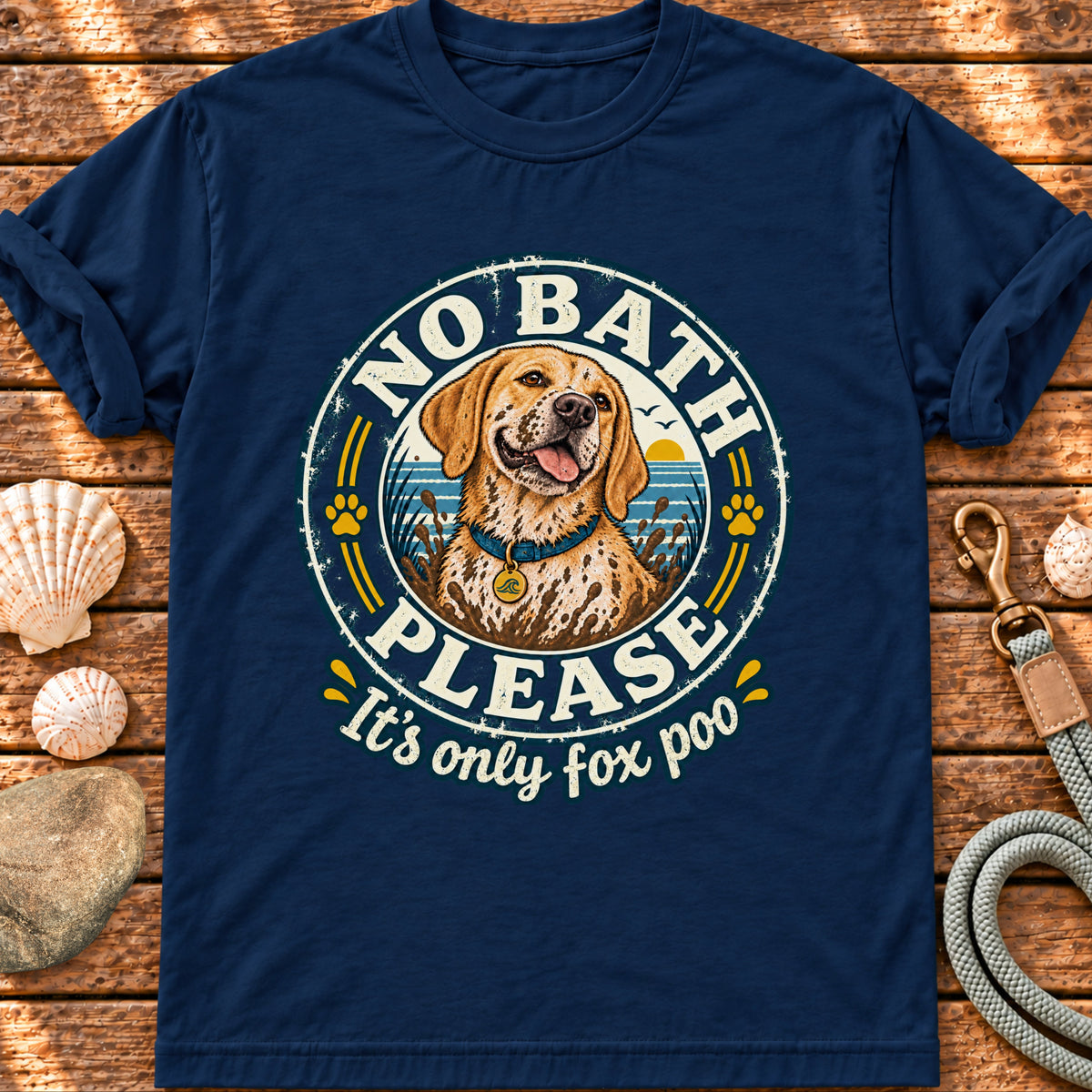 A navy blue short-sleeve t-shirt with a circular graphic of a golden retriever dog and the text 'NO BATH PLEASE It's only fox poo', displayed flat on a rustic wooden background with seashells and a leash.