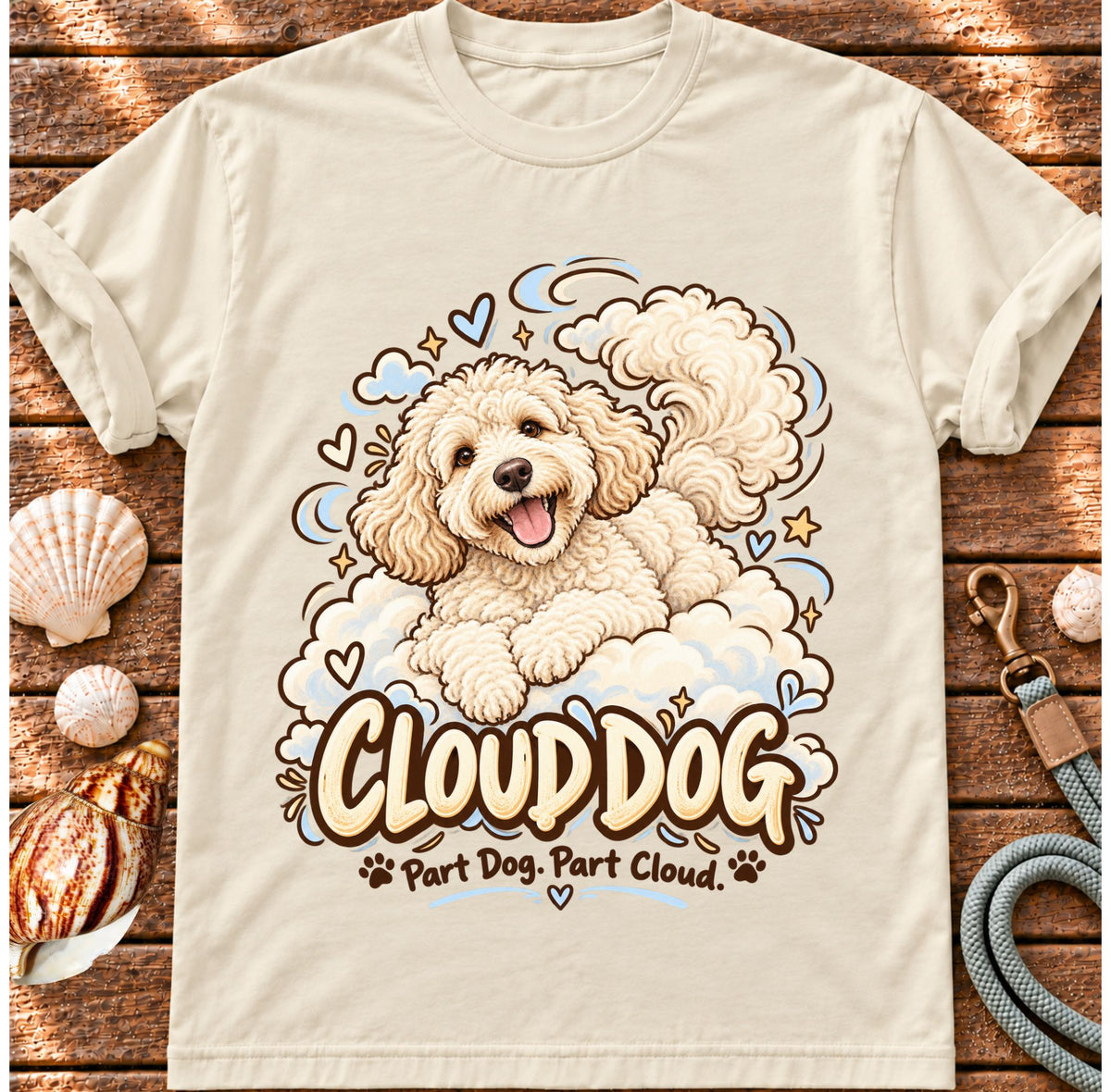 Ivory Cloud Dog T-shirt laid flat on wood, featuring a fluffy white dog surrounded by blue cloud outlines and stars and the wording “Cloud Dog — Part Dog. Part Cloud.”