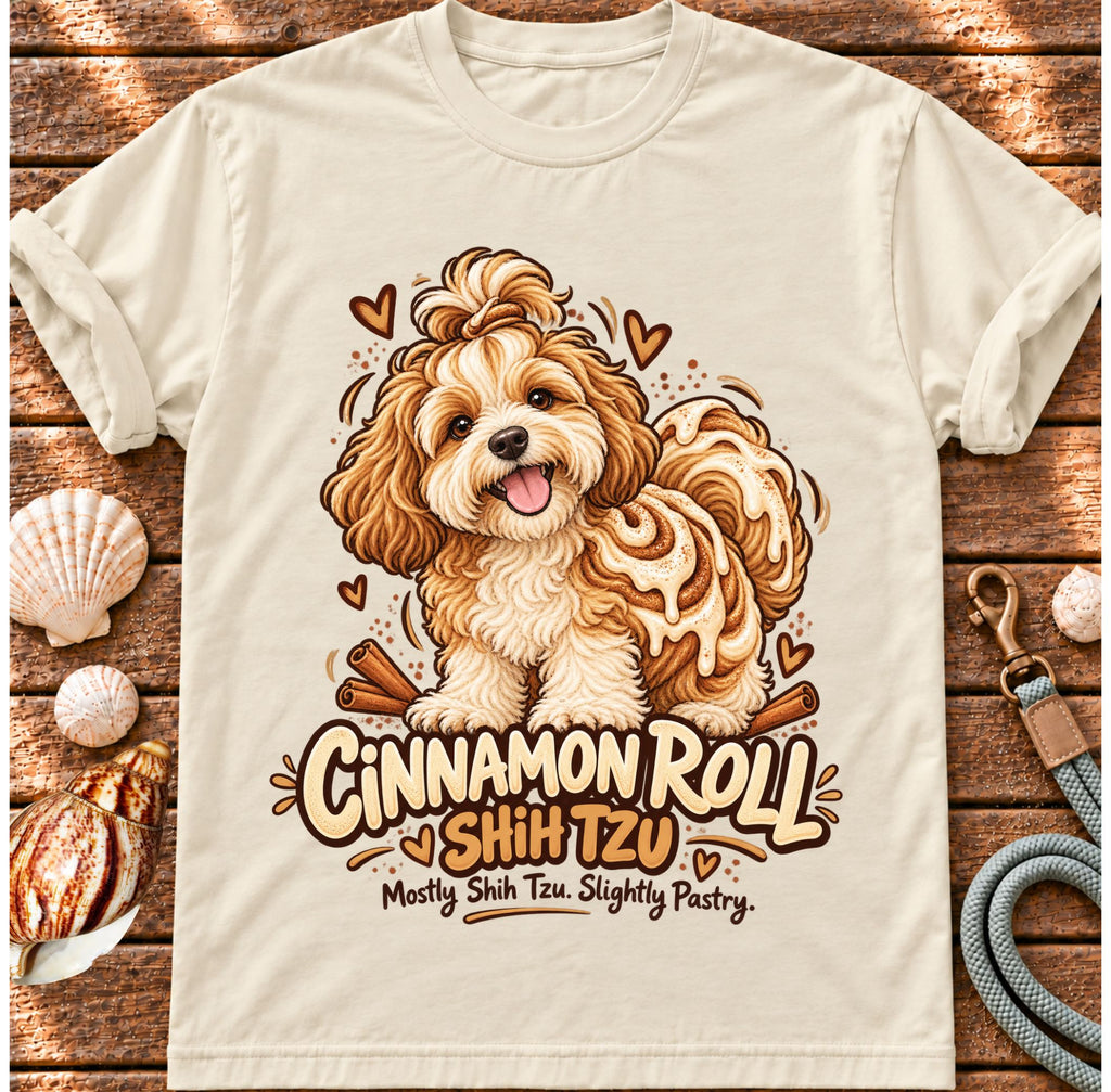 Ivory Cinnamon Roll Shih Tzu T-shirt laid flat on wood, featuring a shaggy Shih Tzu combined with a cinnamon pastry swirl and the wording “Cinnamon Roll Shih Tzu — Mostly Shih Tzu. Slightly Pastry.”