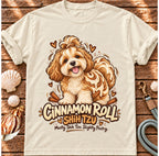 Ivory Cinnamon Roll Shih Tzu T-shirt laid flat on wood, featuring a shaggy Shih Tzu combined with a cinnamon pastry swirl and the wording “Cinnamon Roll Shih Tzu — Mostly Shih Tzu. Slightly Pastry.”
