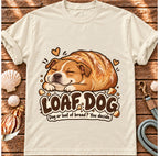 Ivory Loaf Dog T-shirt laid flat on wood, featuring a sleeping dog shaped like a golden loaf of bread and the wording “Loaf Dog — Dog or loaf of bread? You decide.”