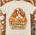 Ivory Croissant Spaniel T-shirt laid flat on wood, featuring a Cavalier spaniel combined with a golden croissant and the wording “Croissant Spaniel — Mostly Spaniel. Slightly Pastry.”