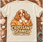 Ivory Croissant Spaniel T-shirt laid flat on wood, featuring a Cavalier spaniel combined with a golden croissant and the wording “Croissant Spaniel — Mostly Spaniel. Slightly Pastry.”