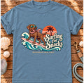 Flat-lay Brinehound T-shirt showing a surfing chocolate Labrador with waves, a sunset and Surfing for Snacks lettering.