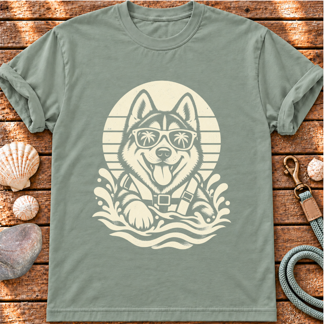 A sage green short-sleeve t-shirt laid flat on a rustic wooden surface, featuring a cream-colored graphic of a husky dog wearing sunglasses and a life vest inside a circular sun design. The shirt is styled with seashells and a leather leash around it.
