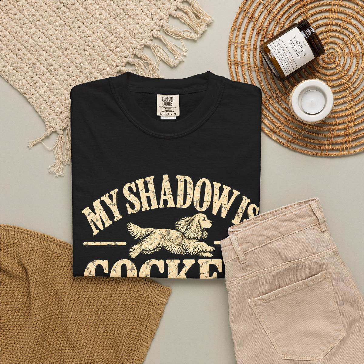 Black T-shirt with the Cocker Spaniel illustration and “My shadow is cocker shaped” lettering, styled with beige trousers and coffee.