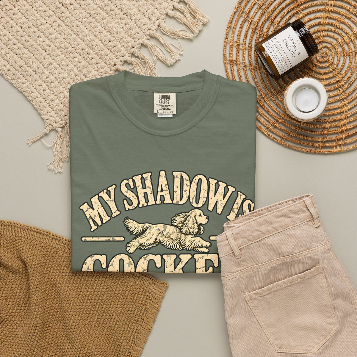 Moss T-shirt with the Cocker Spaniel illustration and “My shadow is cocker shaped” lettering, styled with beige trousers and coffee.