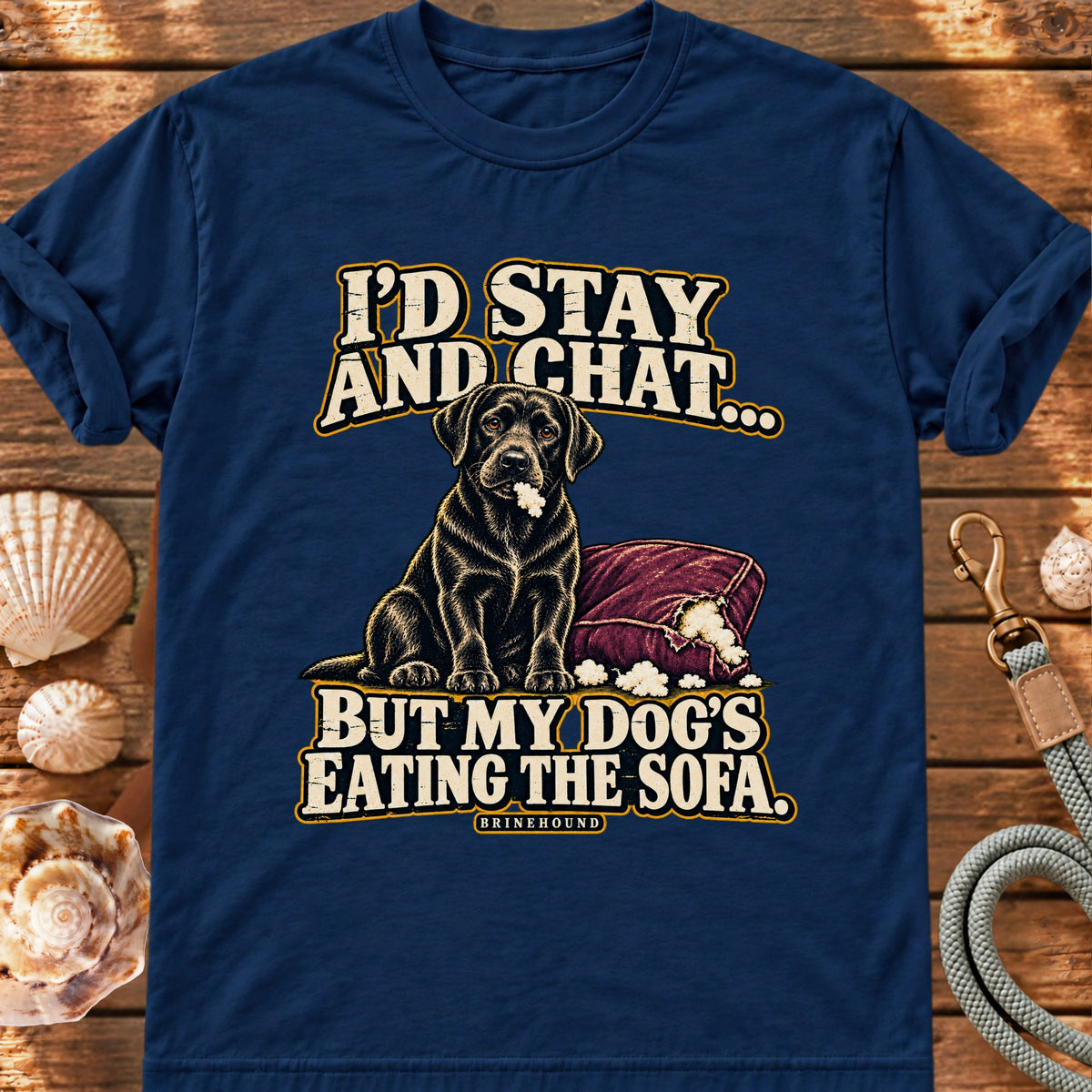 True Navy T-shirt with the black Labrador beside a chewed sofa and “I’d stay and chat” lettering print, laid on wood beside seashells and a dog lead.