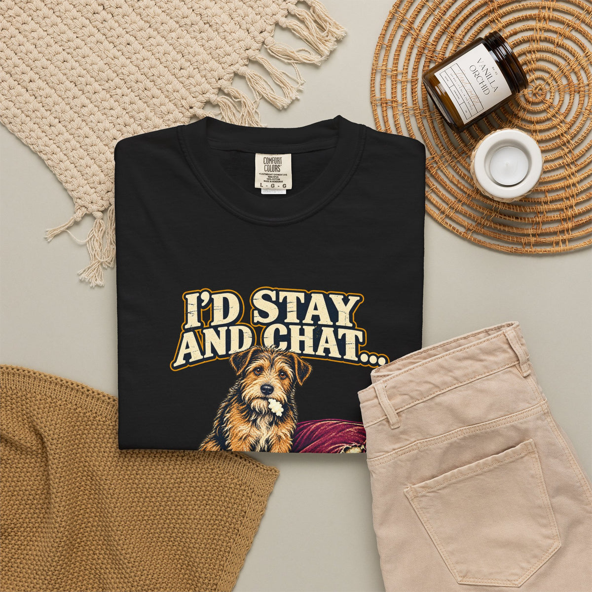 Black T-shirt with the scruffy terrier beside a chewed sofa and “I’d stay and chat” lettering print, styled with beige trousers and coffee.