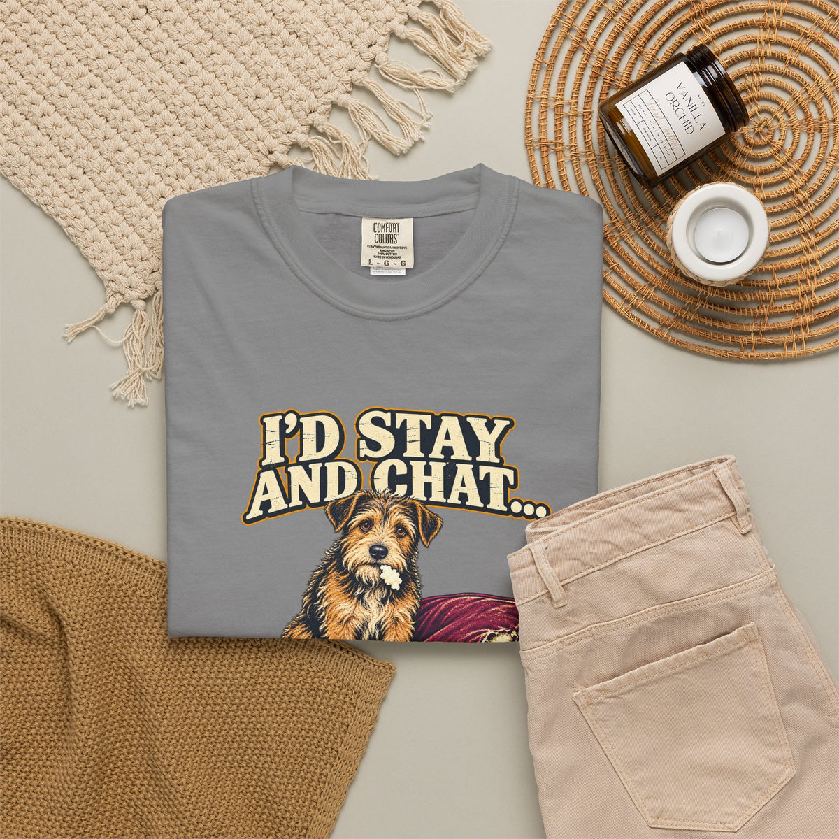Grey T-shirt with the scruffy terrier beside a chewed sofa and “I’d stay and chat” lettering print, styled with beige trousers and coffee.
