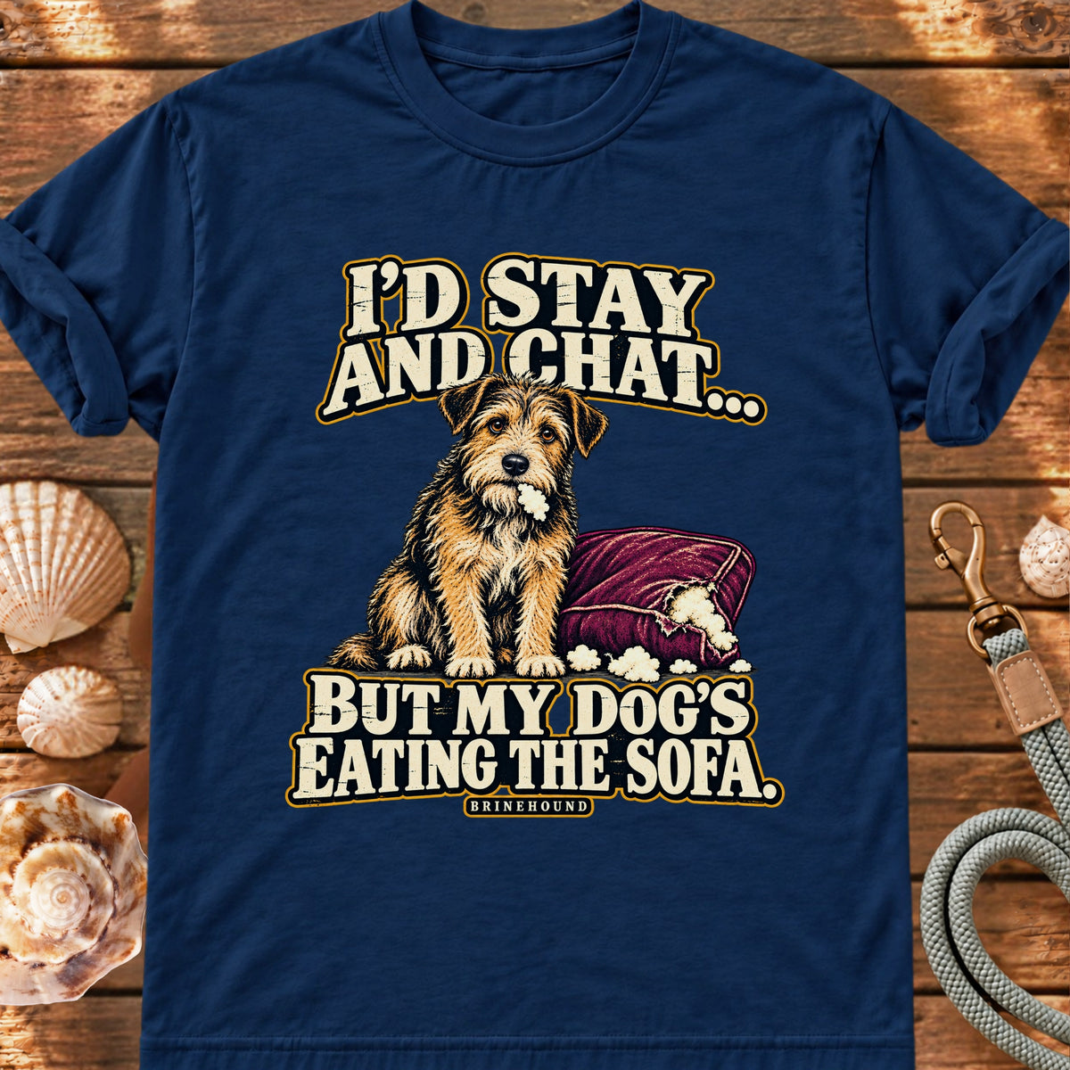 True Navy T-shirt with the scruffy terrier beside a chewed sofa and “I’d stay and chat” lettering print, laid on wood beside seashells and a dog lead.